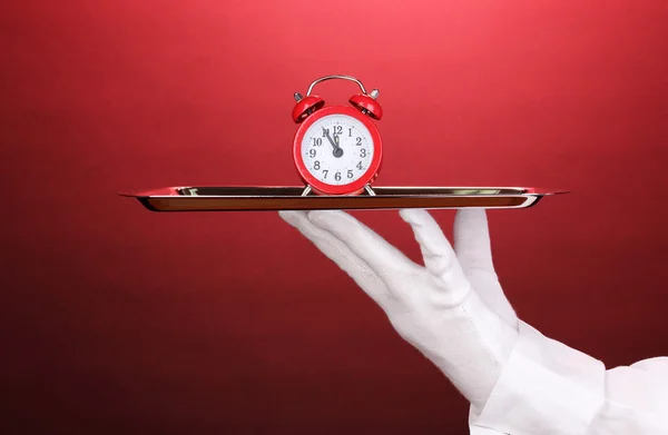 Hand in glove holding silver tray with alarm clock on red background — Stock Photo, Image