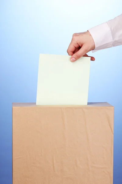 Mano con papeleta de votación y caja sobre fondo azul — Foto de Stock