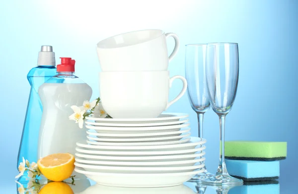 Farfurii curate goale, pahare și căni cu lichid de spălat vase, bureți și lămâie pe fundal albastru — Fotografie, imagine de stoc