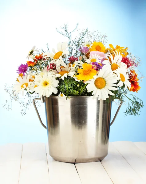 Beautiful bouquet of bright wildflowers in metal bucket, on wooden table on blue background — Stock Photo, Image