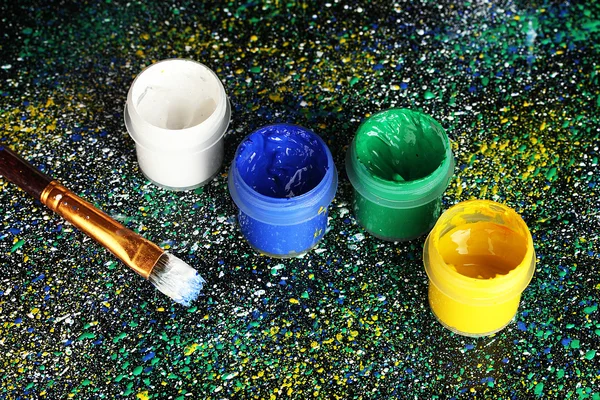 Jars with colorful gouache and brush on black background, spattered with colorful paint close-up — Stock Photo, Image