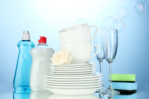 Piatti puliti vuoti, bicchieri e tazze con liquido per piatti e spugne su fondo blu — Foto Stock
