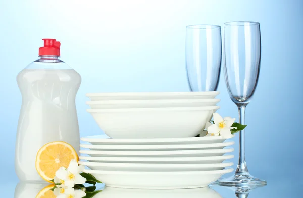 Platos y vasos limpios vacíos con líquido lavavajillas y limón sobre fondo azul —  Fotos de Stock