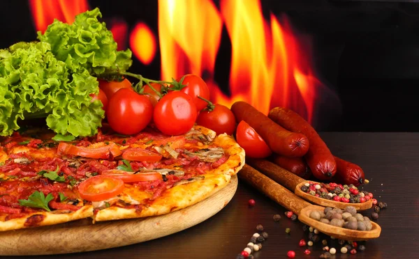 Delicious pizza, salami, tomatoes and spices on wooden table on flame background — Stock Photo, Image