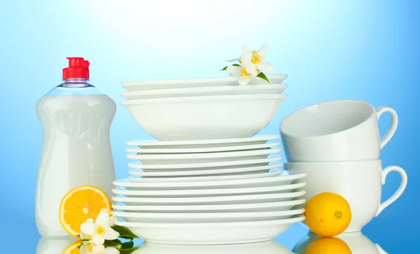 Platos y tazas limpias vacías con líquido lavavajillas y limón sobre fondo azul —  Fotos de Stock