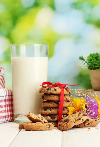 Стакан молока, шоколадное печенье с красной лентой и полевые цветы на деревянном столе на зеленом фоне — стоковое фото