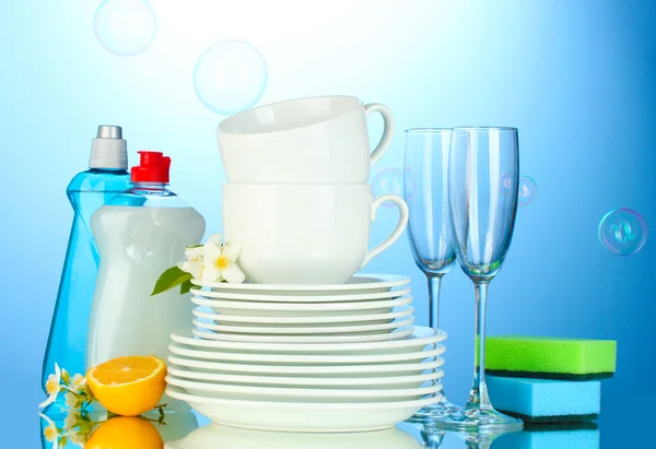 Platos limpios vacíos, vasos y tazas con líquido lavavajillas, esponjas y limón sobre fondo azul — Foto de Stock