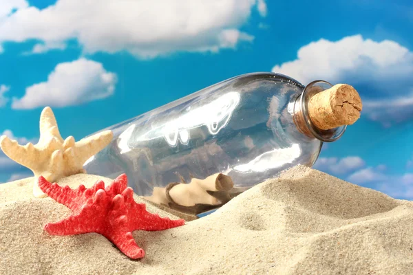 Glazen flesje met opmerking binnen op zand, op blauwe hemelachtergrond — Stockfoto