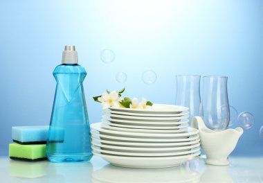 Empty clean plates and glasses with dishwashing liquid, sponges and flowers on blue background clipart