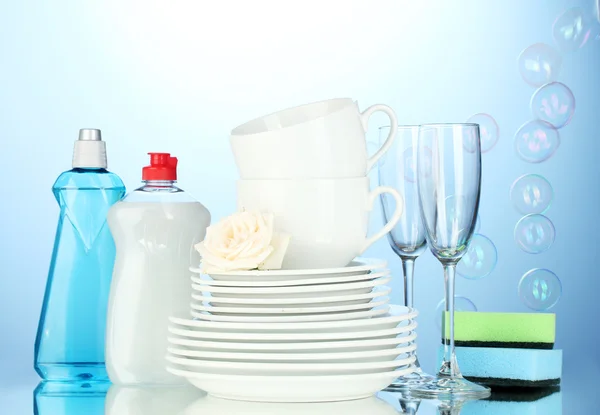 Пустые чистые тарелки, стаканы и чашки с жидкостью для мытья посуды и губки на синем фоне — стоковое фото