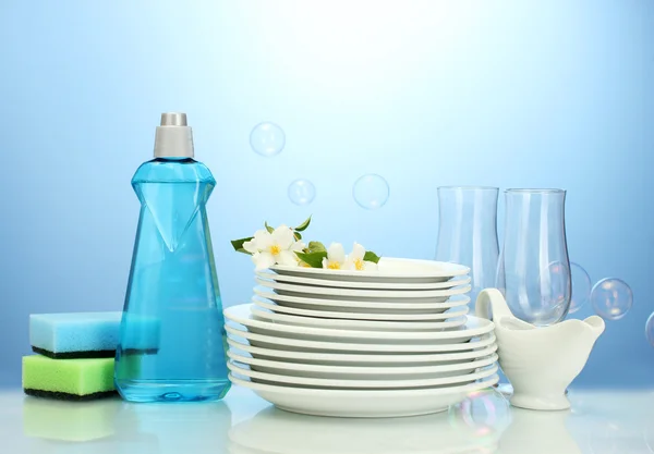 Platos y vasos limpios vacíos con líquido lavavajillas, esponjas y flores sobre fondo azul — Foto de Stock