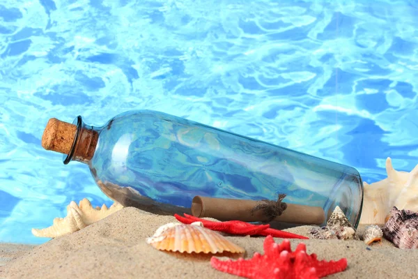 Glass bottle with note inside on sand, on blue sea background — Stock Photo, Image