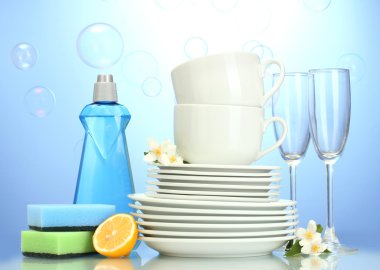 Empty clean plates, glasses and cups with dishwashing liquid, sponges and lemon on blue background clipart