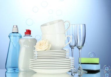 Empty clean plates, glasses and cups with dishwashing liquid and sponges on blue background clipart