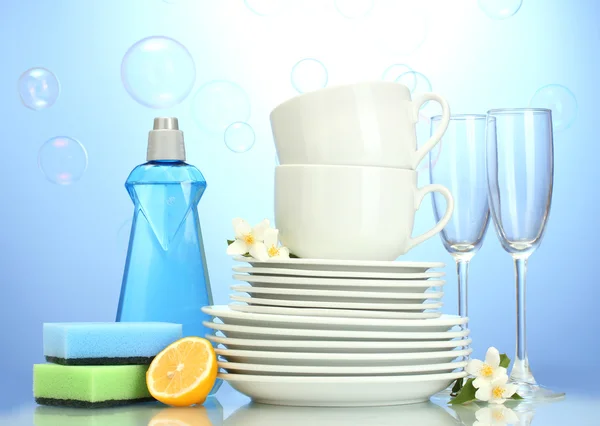 Assiettes propres vides, verres et tasses avec liquide vaisselle, éponges et citron sur fond bleu — Photo