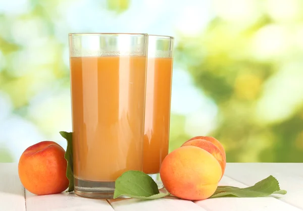 Glasses of apricot juice and fresh apricots on white wooden table on green background — Stock Photo, Image