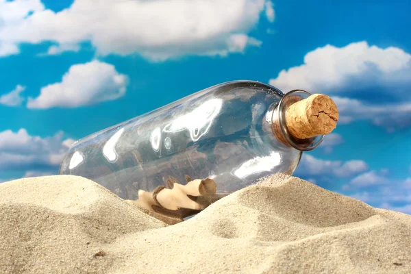 Glazen flesje met opmerking binnen op zand, op blauwe hemelachtergrond — Stockfoto