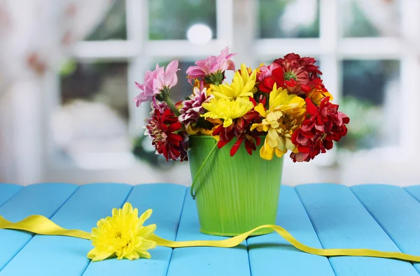Zielony wiaderko z kwiatami na niebieskim drewnianym stole na tle okna — Zdjęcie stockowe