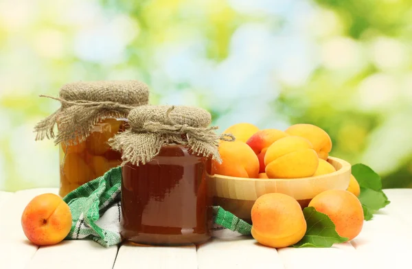 Canned apricots and jam in a jars and ripe apricots in bowl on wooden table on green background — Stock Photo, Image