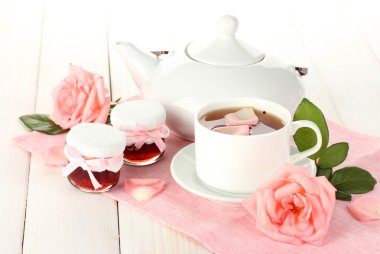 Teapot and cup of tea with roses and jam on white wooden table clipart