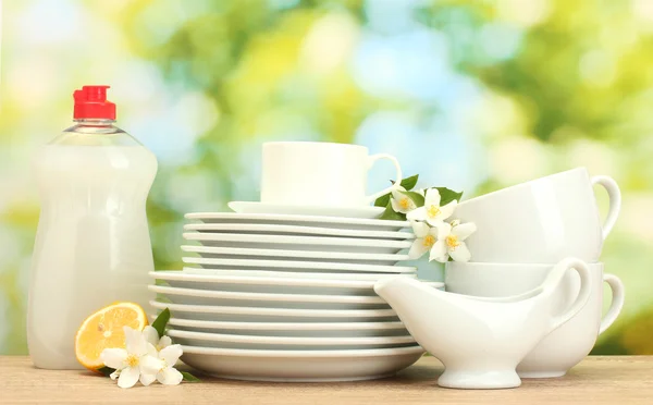 Pusty czyste talerze i kubki z płynu do mycia naczyń, kwiaty i cytryny na drewnianym stole na zielonym tle — Zdjęcie stockowe