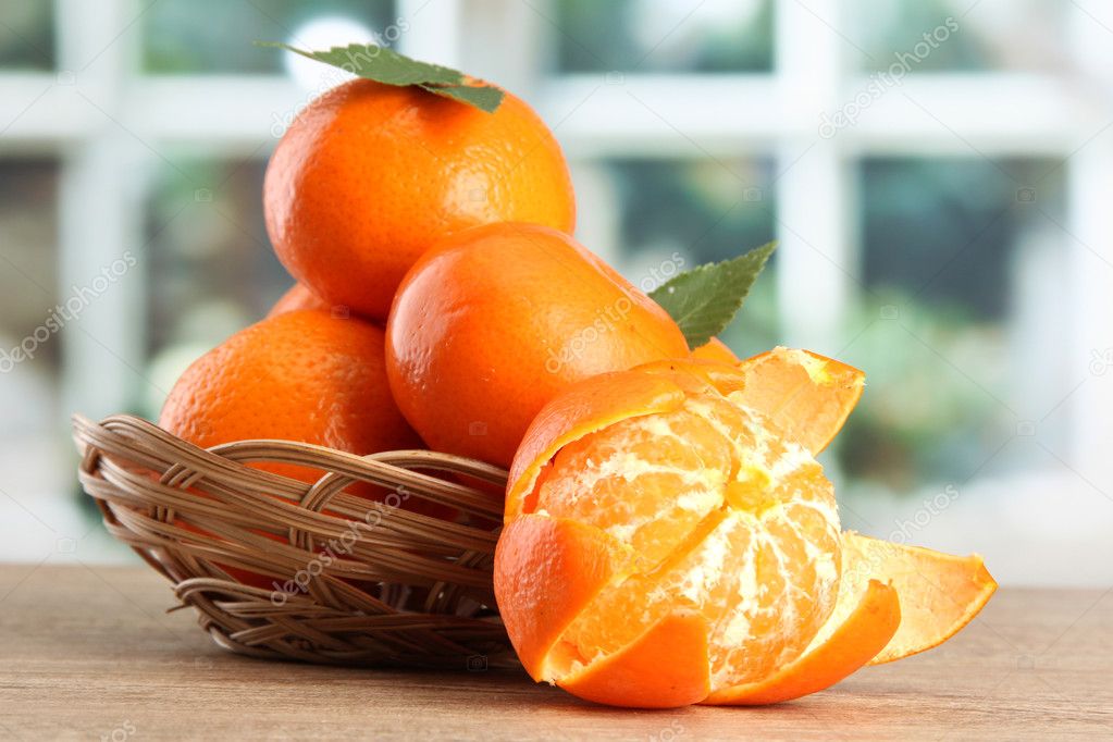 Tangerines with leaves in a beautiful basket, on wooden table on window background