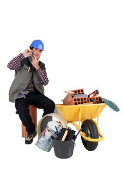 Mans sat making call next to wheelbarrow full of bricks clipart