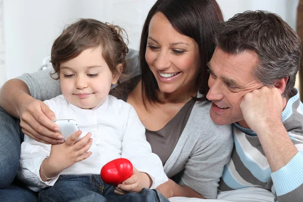Kleinkind hält Handy in der Hand, während es mit Eltern auf dem Sofa sitzt — Stockfoto