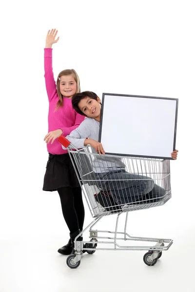 Children in a shopping trolley with a blank board — Stock Photo, Image