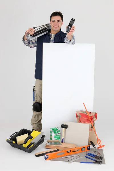 Tradesman holding a mobile phone over a blank sign and his tools — Stock Photo, Image