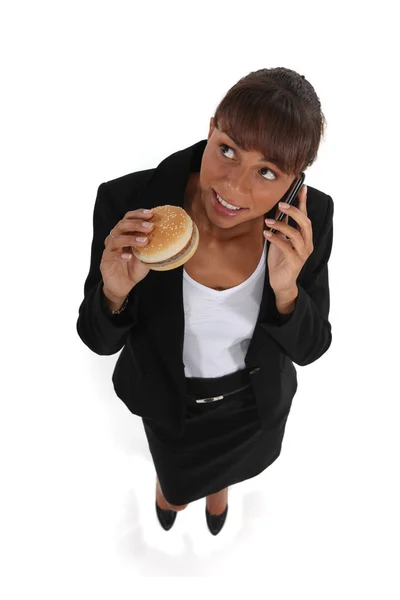 Businesswoman eating a hamburger and talking on her cell — Stock Photo, Image