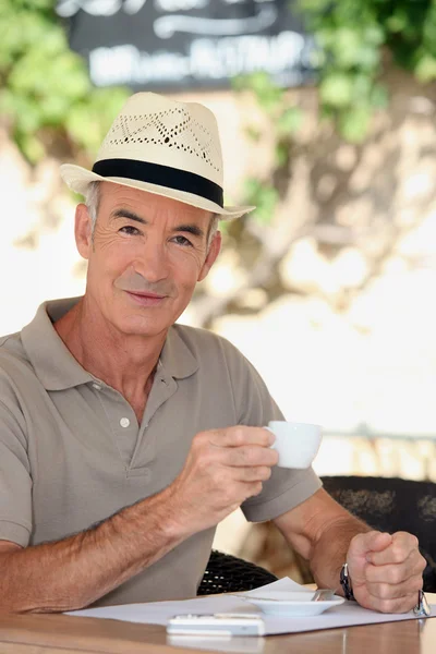 65 år gammal man klädd i en stråhatt och dricka en kopp te på ett kafé terrass — Stockfoto