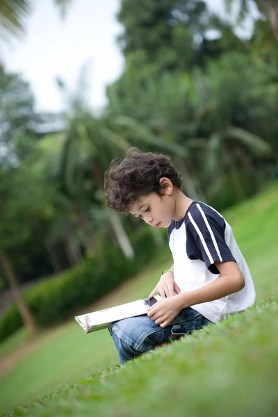Mladý chlapec těší jeho čtení knihy ve venkovním parku — Stock fotografie