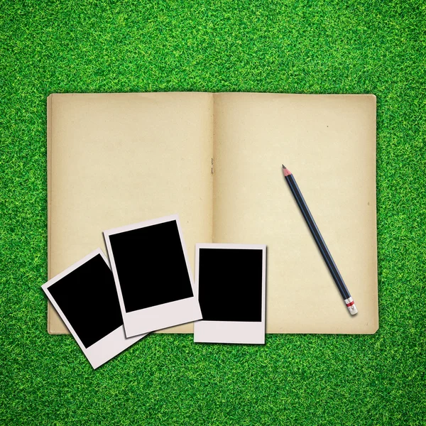 Pencil and photo frame with old book on green grass background — Stock Photo, Image