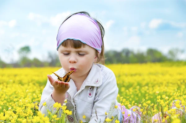 Lilla söta tjejen i ett fält av vackra gula blommor håller en fjäril på handflatorna och blåser på den. Royaltyfria Stockfoton