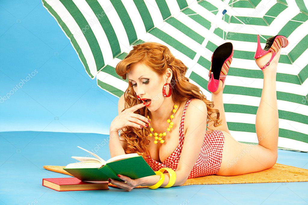 Beautiful sexy girl pin up in a pink bikini reads book on the beach under sun umbrella