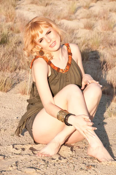 Beautiful blonde in a khaki shirt sitting cross-legged on sand — Stock Photo, Image