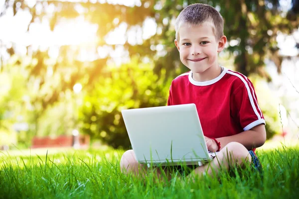 Milý chlapeček sedí s notebookem v trávě — Stock fotografie