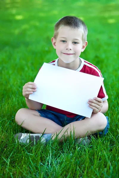 Menino no prado verde com uma folha de papel branca — Fotografia de Stock