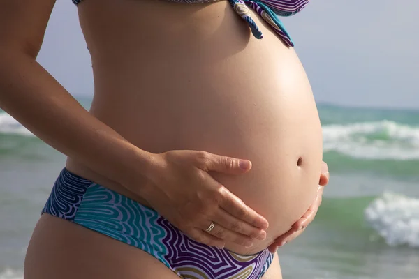 Закрытие рук, держащих большой беременный живот перед морем — стоковое фото