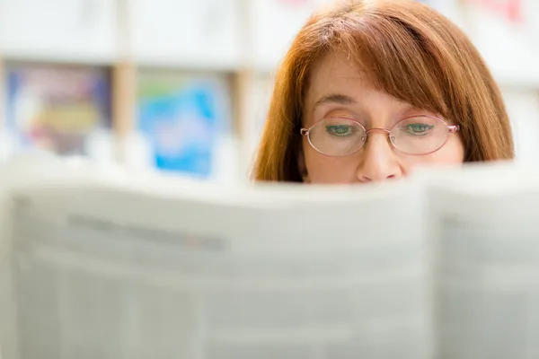 Mulher idosa com óculos lendo jornal na biblioteca — Fotografia de Stock