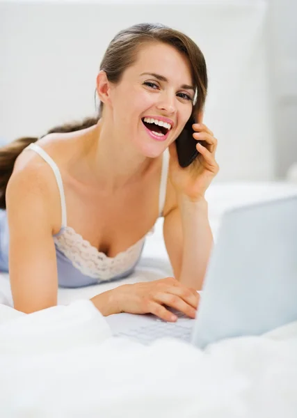 Χαμογελαστή γυναίκα τοποθέτηση στο κρεβάτι με το lap-top, πραγματοποίηση τηλεφωνικής κλήσης — Φωτογραφία Αρχείου