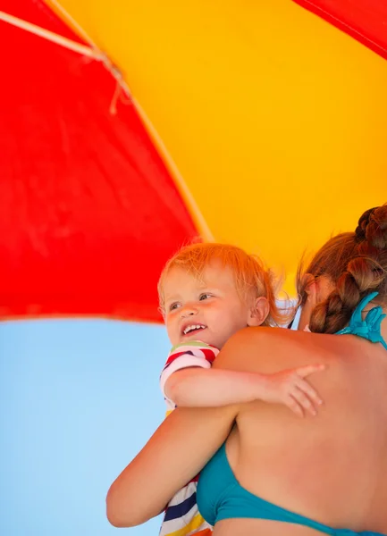 Mãe segurando bebê olhando no espaço cópia na praia sob guarda-chuva — Fotografia de Stock