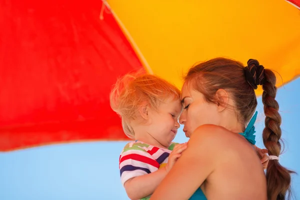 Moeder zoenen baby op strand onder de paraplu — Stockfoto