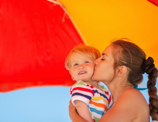 Мать целует ребенка на пляже под зонтиком — стоковое фото