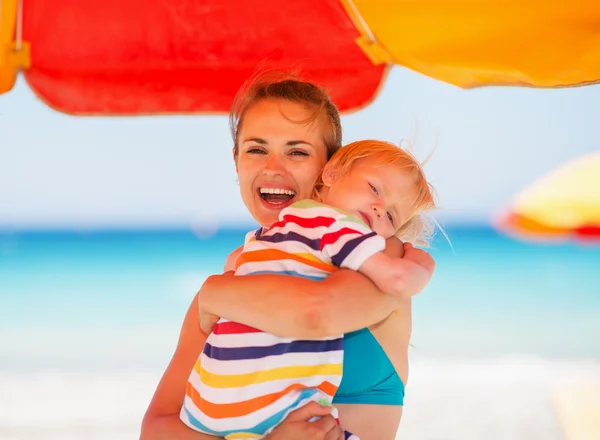 Mother embracing baby on beach under umbrella — Stock Photo, Image