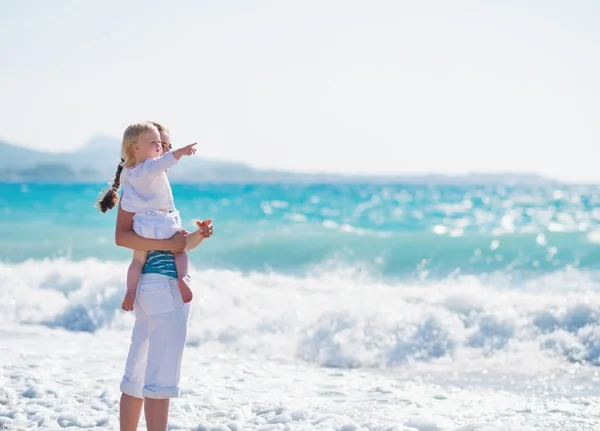 Moeder aan zee oever met baby wijzen op kopie ruimte — Stockfoto