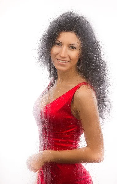 Hermosa chica en un vestido rojo, de pie detrás de un vaso mojado sobre un fondo blanco . — Foto de Stock