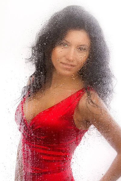 Bella ragazza in un abito rosso, in piedi dietro un vetro bagnato su uno sfondo bianco . — Foto Stock