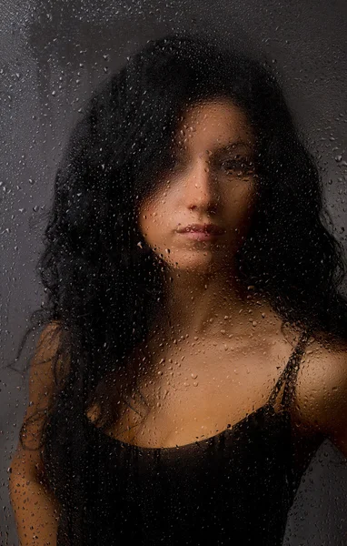 Красивая девушка за мокрым стеклом на темном фоне . — стоковое фото